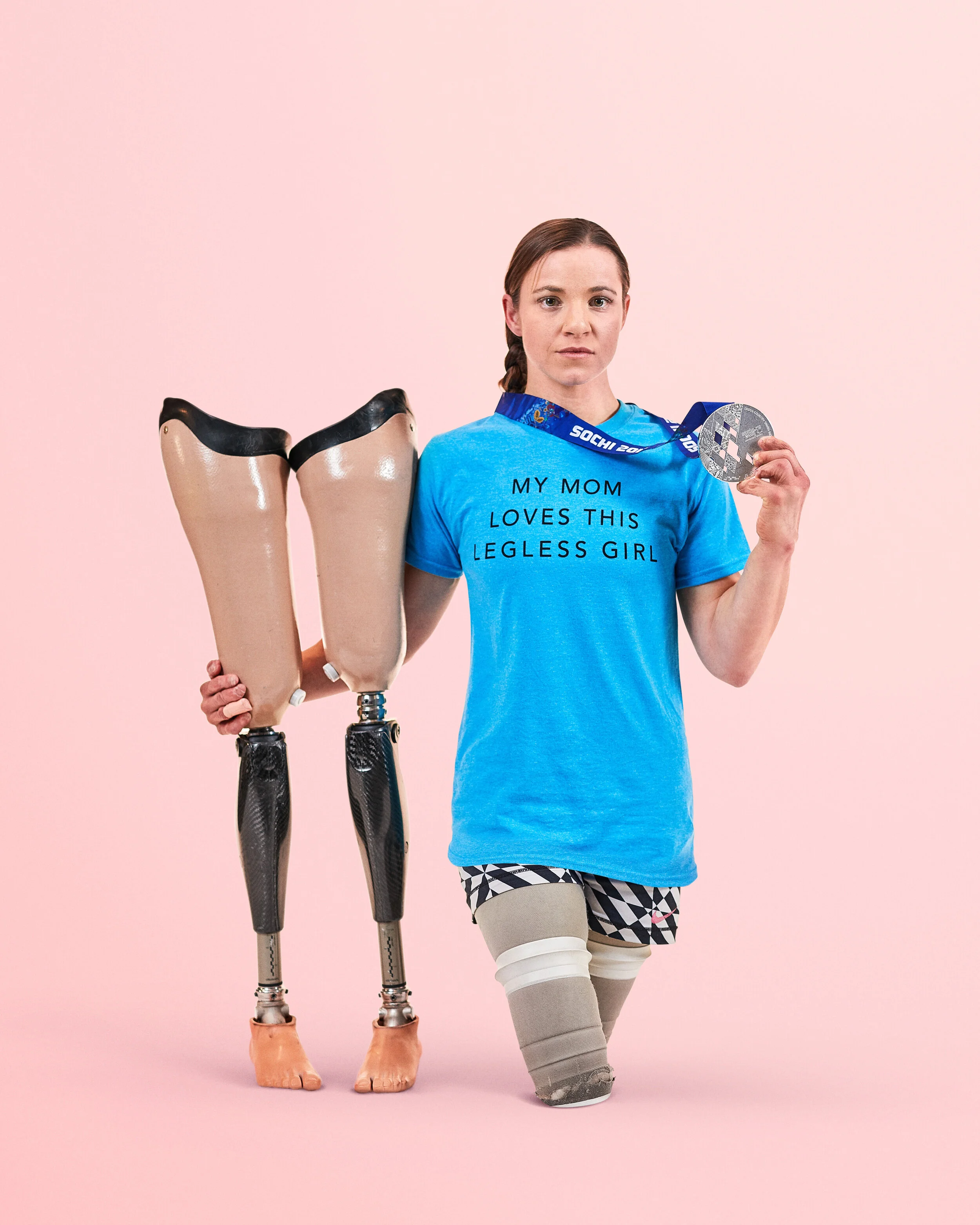 Amy Purdy holding prosthetic legs and wearing a blue campaign T-shirt against a pink background