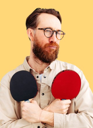 A smiling woman with her arms crossed, standing against a dark green background. She has long, dark hair.