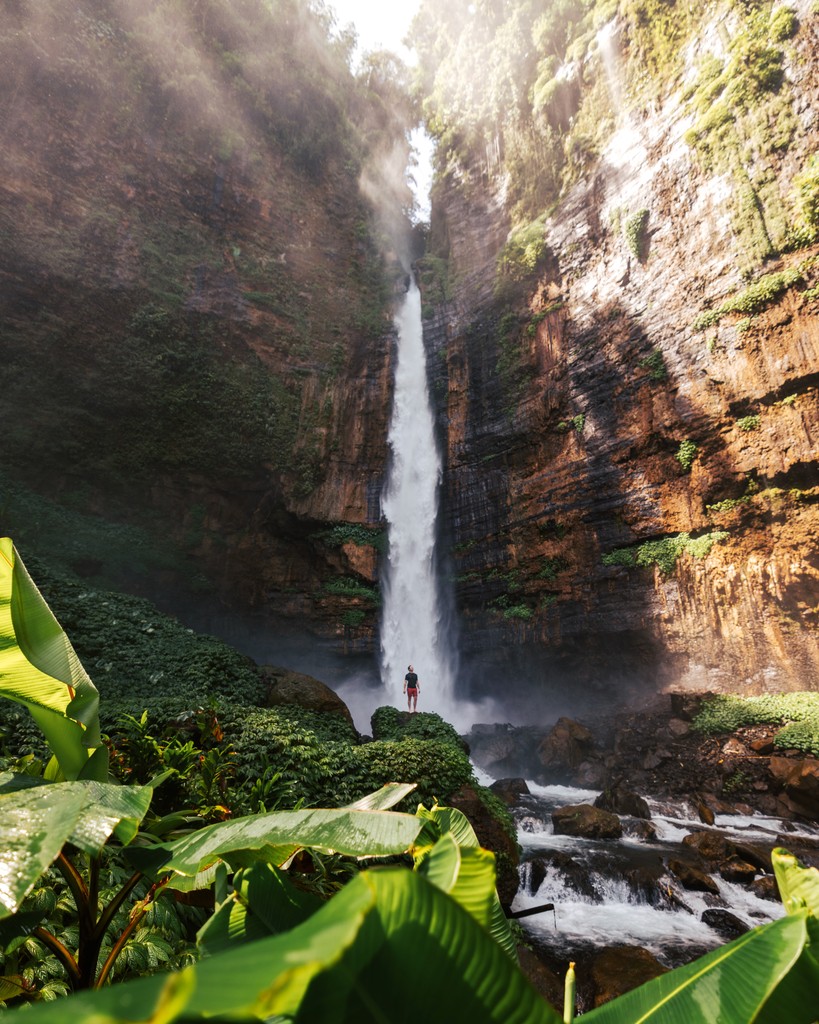 A man standing on a leaf-covered rock looking up at the rock formation above with a waterfall in the background