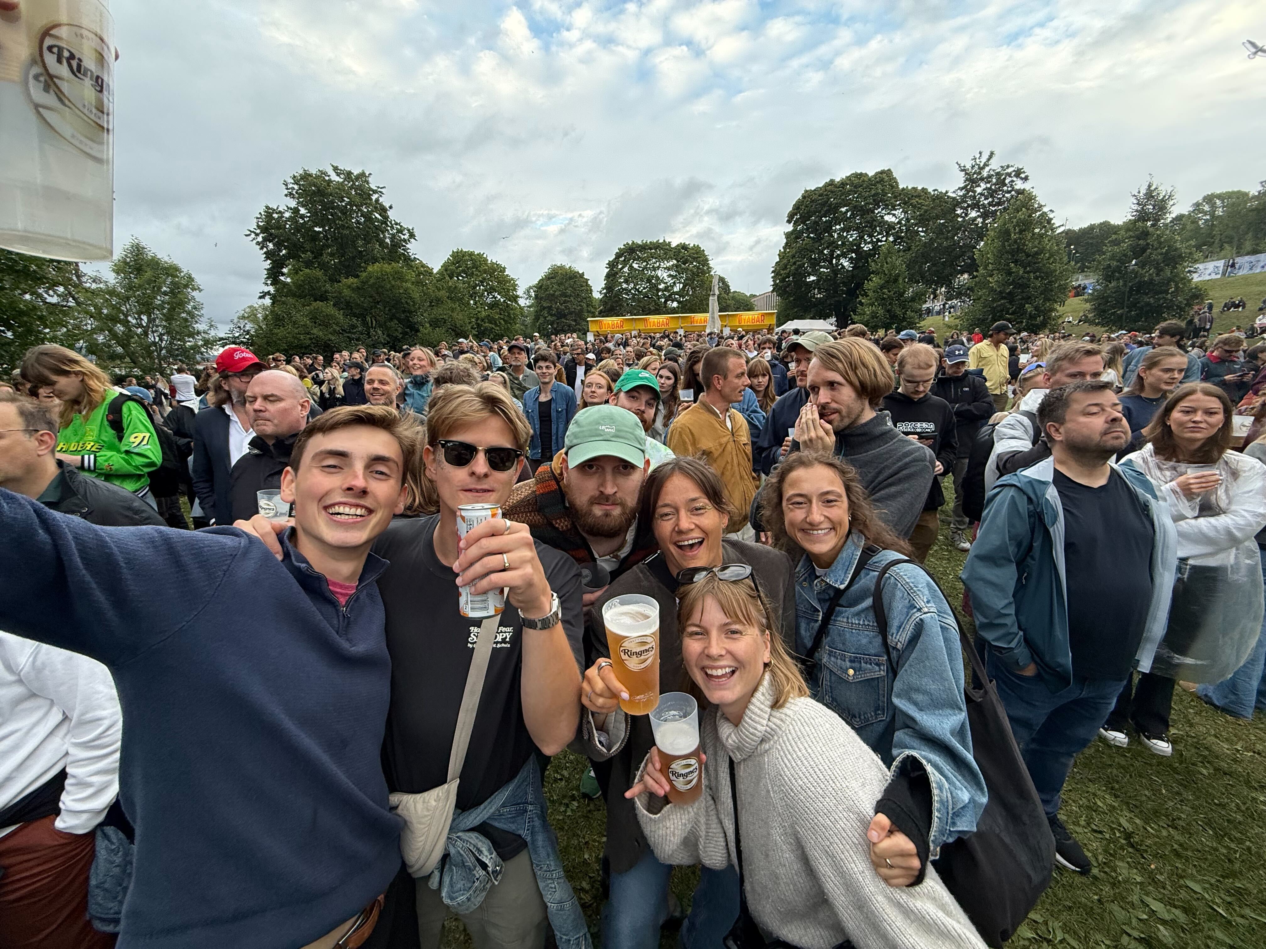 Pictures of a group of people smiling and cheering towards the camera. Located at a festival arena with a crowd in the background.