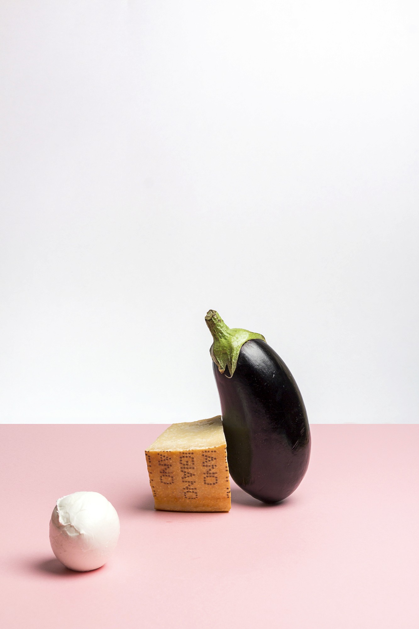 Nature morte studio sur une table au fond rose d'ingrédients mis en scène comme une aubergine, un morceau de parmesan et une boule de mozarella