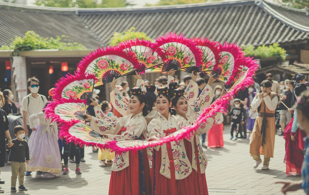 A group of performers dressed in traditional Korean hanbok outfits execute a beautiful fan dance with vibrant, pink-feathered fans during a cultural festival, captivating the crowd with their graceful movements.