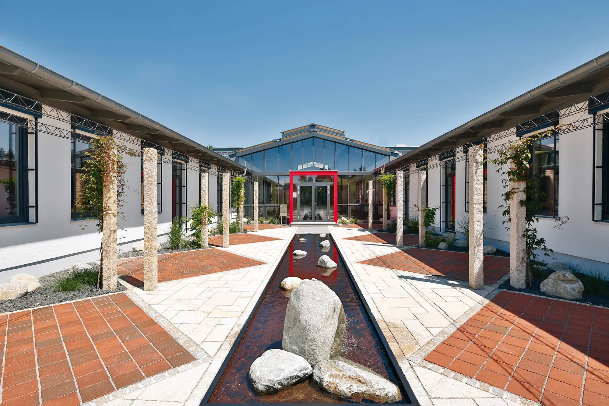 Moderne Architektur mit Wasserspielanlage und roten Ziegelsteinen auf dem Boden, umgeben von gepflegten grünen Pflanzen.