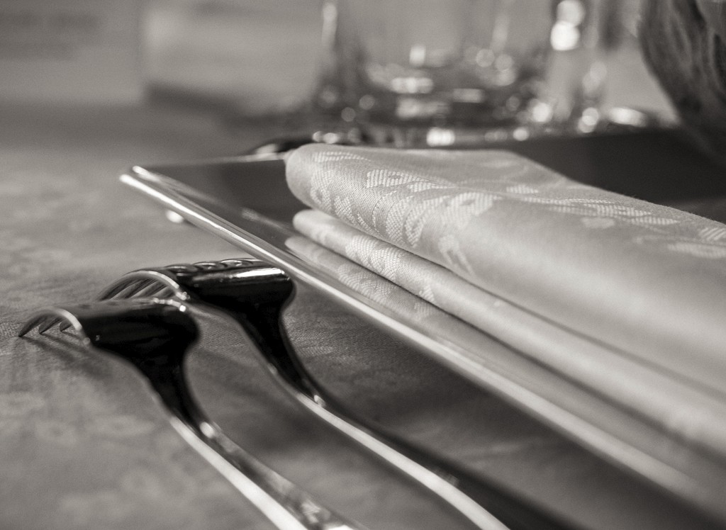 Fine art close-up of elegant wedding tableware — folded linen napkin and polished silver cutlery reflecting soft ambient light. Détail artistique d’une table de mariage raffinée — serviette en lin pliée et couverts en argent poli reflétant une lumière douce et chaleureuse.