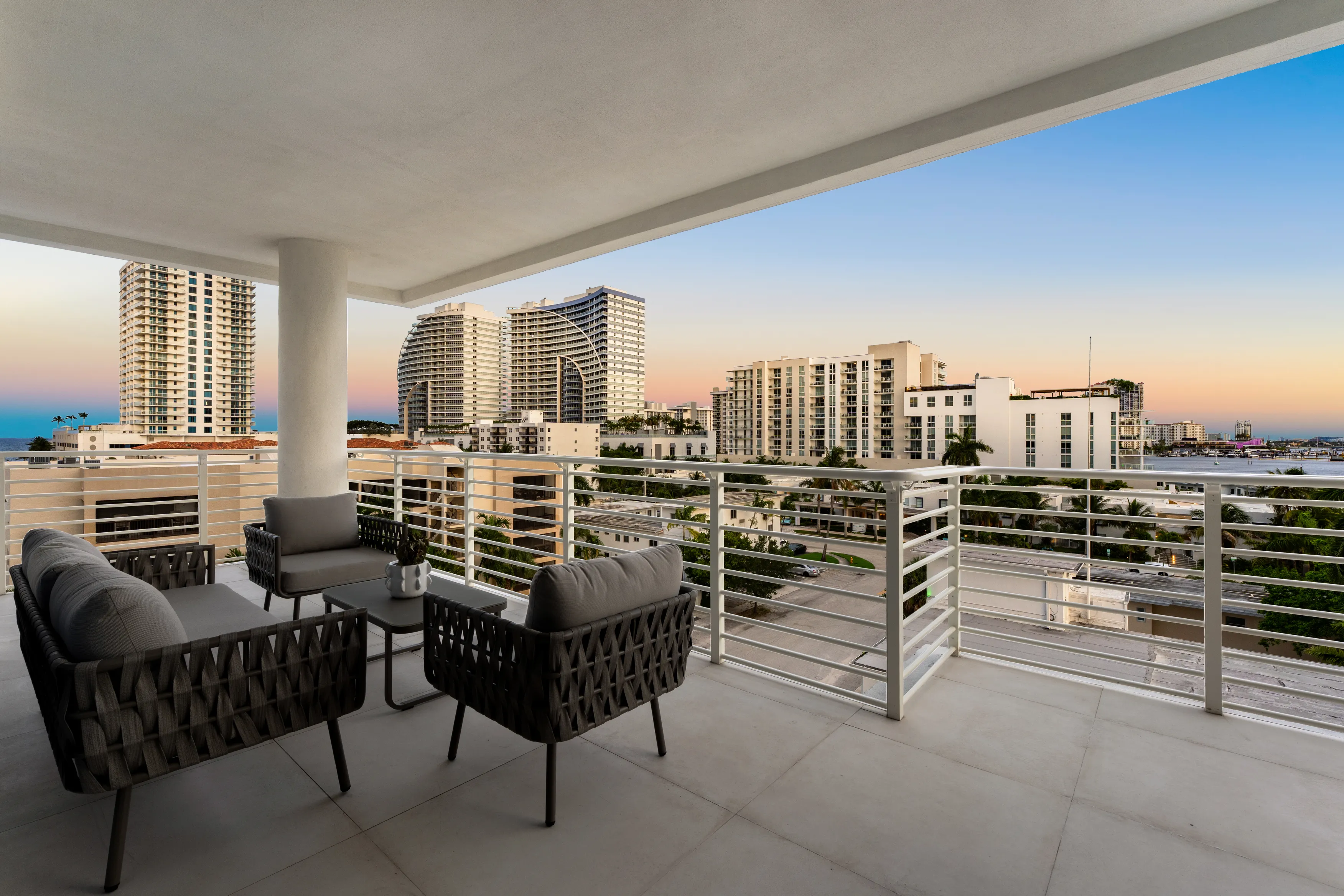 Modern apartment balcony with outdoor dining area, glass doors leading to a living room, city buildings, and greenery visible in the background.
