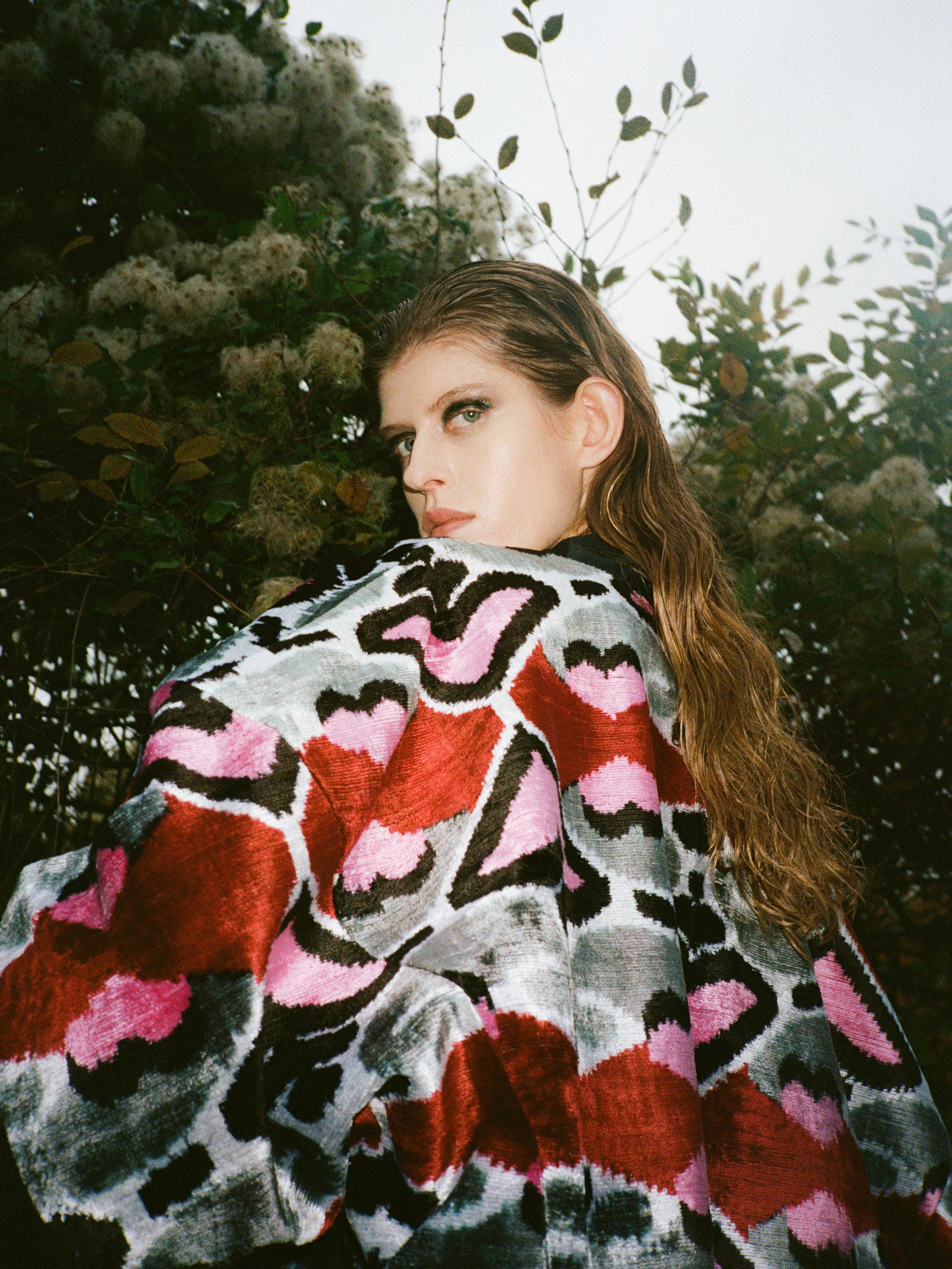 Woman with long hair looking over her shoulder, wearing a patterned sweater with pink, red, black, and white abstract designs.