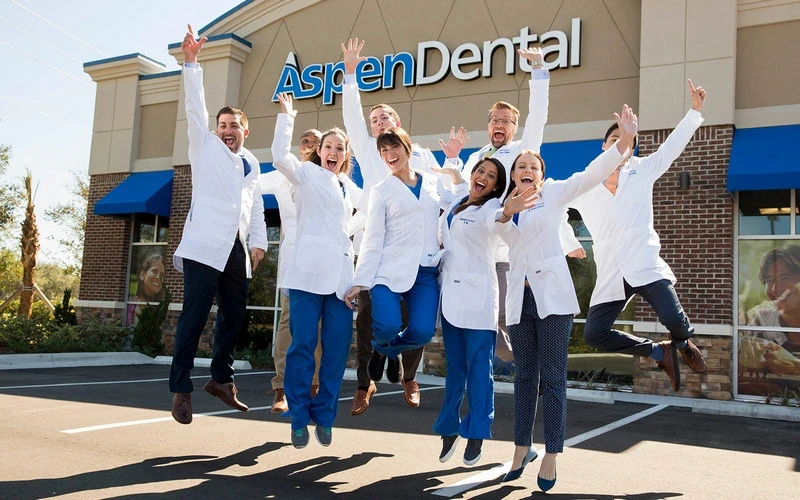 Group of Aspen Dental staff members in white coats jumping with excitement outside an Aspen Dental clinic, celebrating in front of the building’s signage. Captures a positive and energetic team culture focused on patient care.