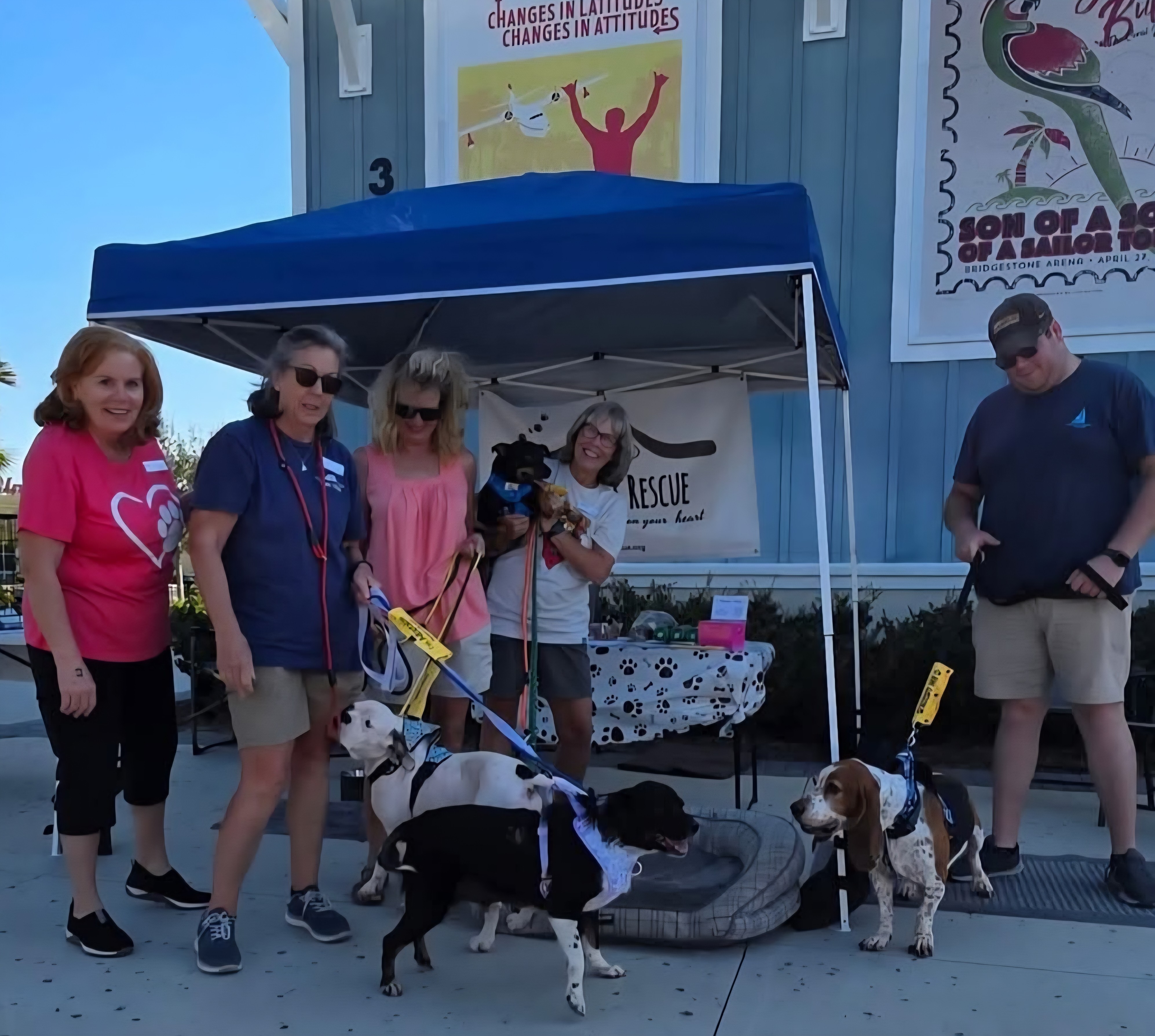 A group of people stand with several dogs in front of a blue tent at an outdoor event.