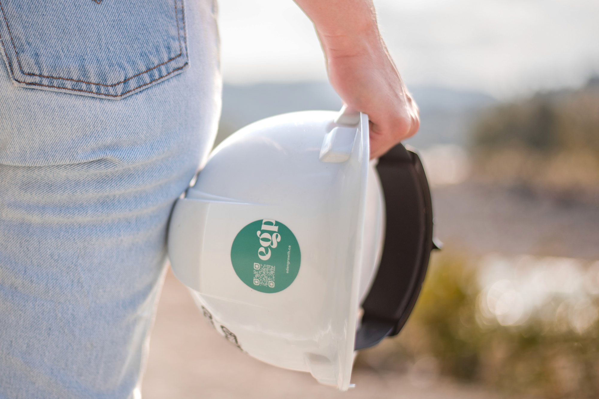 Close-up of a white hard hat with a green logo sticker held against a pair of jeans