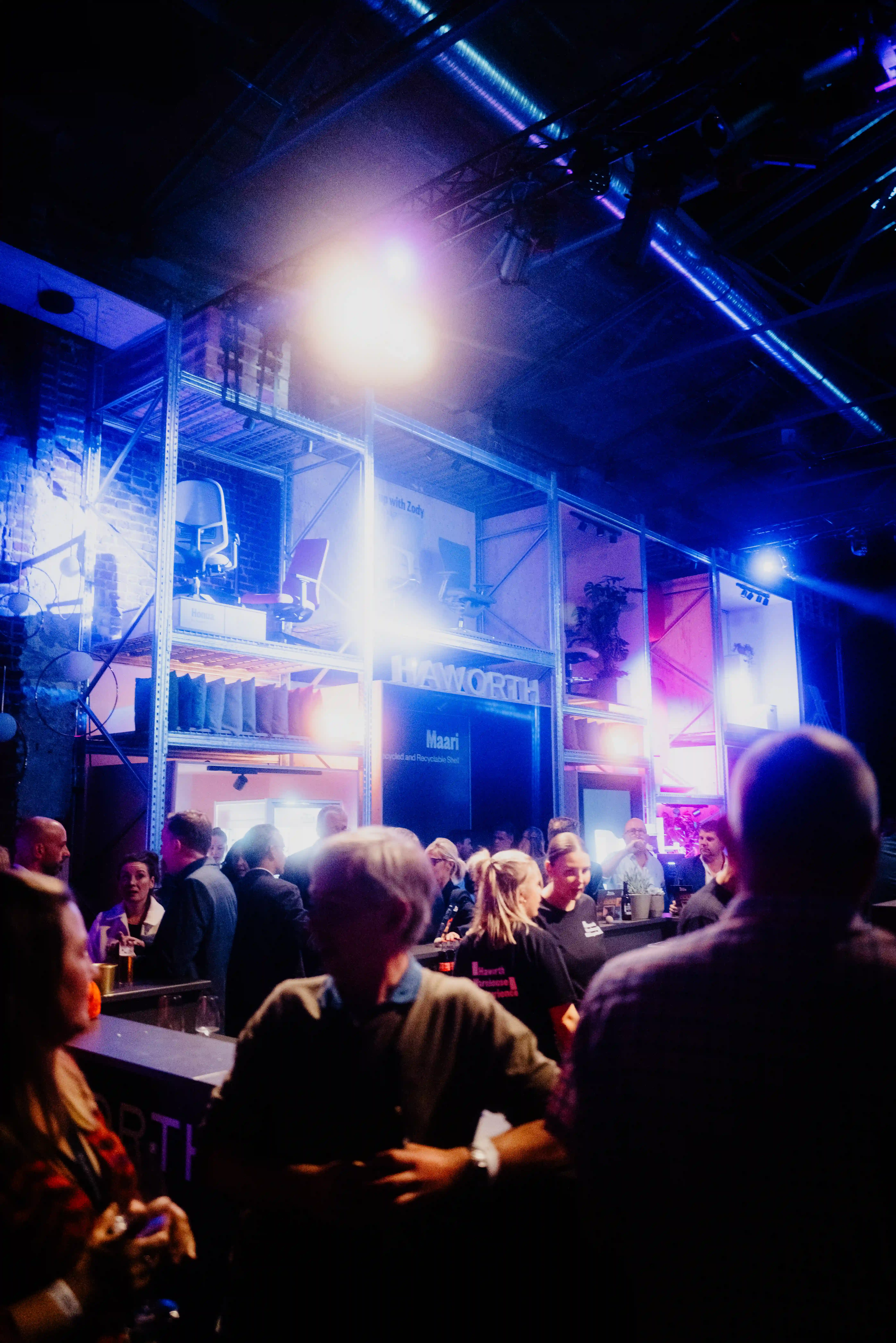 Evening crowd at the bar of the Haworth booth at ORGATEC, with the illuminated shelving structure glowing in blue light.
