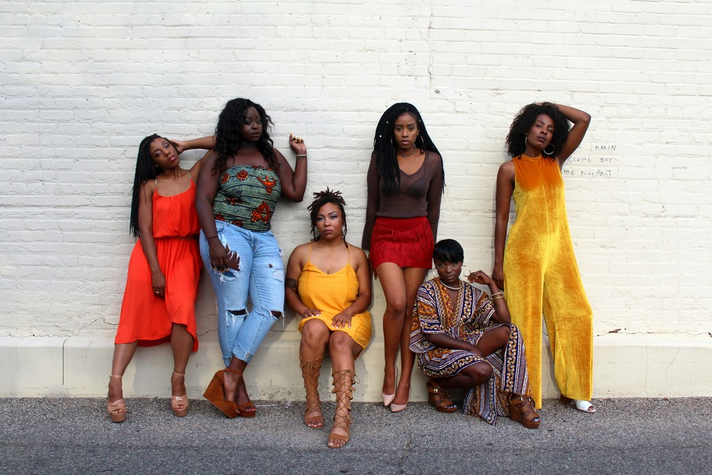 A group of stylish women pose confidently against a white brick wall, showcasing a diverse range of vibrant, fashionable outfits and exuding a strong sense of individuality and empowerment.