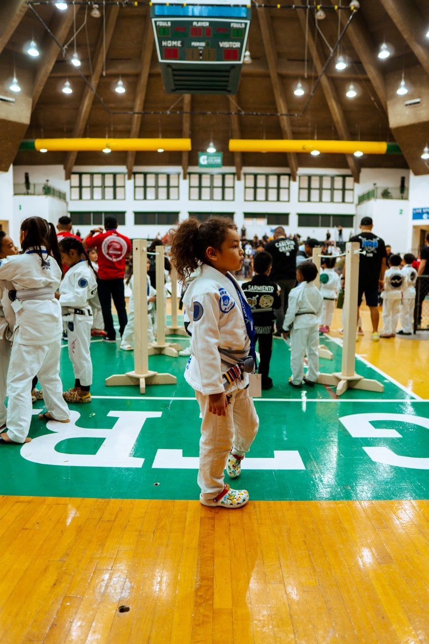 A young girl in a white jiu-jitsu uniform stands on the side of the mat, observing a Pacific Grappling Events tournament, with other children and spectators in the background.