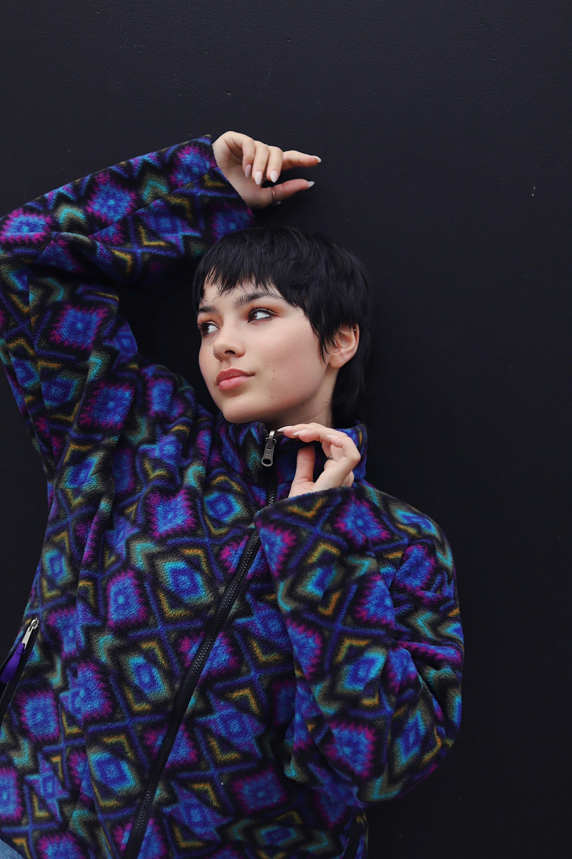 A person with short, dark hair and a colorful patterned fleece jacket posing against a black background with one arm raised.