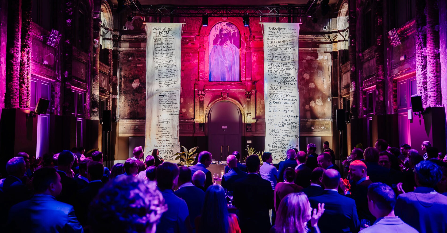 Publiek tijdens evenement in historisch verlichte zaal met grote beschilderde doeken en paarse verlichting.