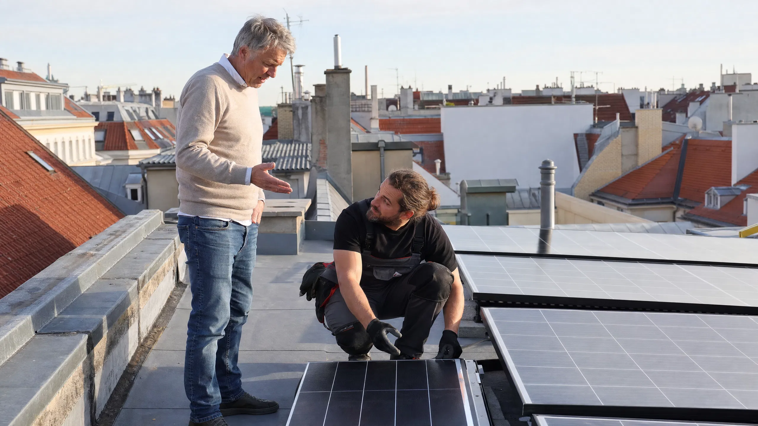 Ein Vermieter spricht auf dem Flachdach eines Wiener Zinshauses mit einem Solartechniker, der kniend eine neu installierte Photovoltaikanlage überprüft, im Hintergrund die typische Wiener Dachlandschaft.