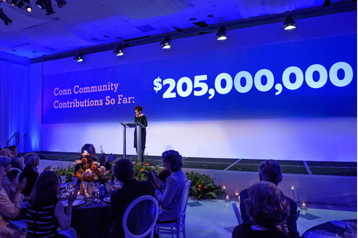 Woman at podium with blue screen behind that says "Conn community contributions so far: $205,000,000"