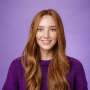 Smiling woman with long, wavy hair wearing a purple sweater against a purple background.