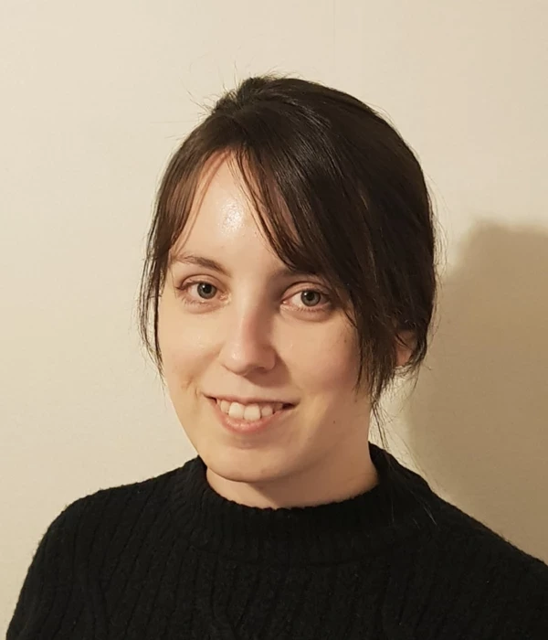 A portrait‑style photograph of a person shown from the shoulders up against a plain, cream‑coloured wall. They are wearing a black knitted jumper with a subtle textured pattern. Dark brown hair is parted to one side and pulled back neatly, with a few loose strands visible around the ear. The lighting is soft and warm, creating a simple, understated, and professional head‑and‑shoulders portrait.