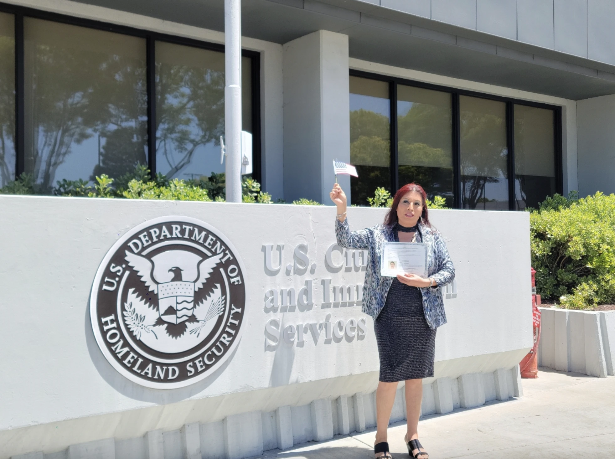 Uma pessoa está orgulhosamente em frente ao prédio do Serviço de Cidadania e Imigração dos EUA, segurando uma pequena bandeira americana e um certificado, celebrando uma conquista significativa com um traje profissional.