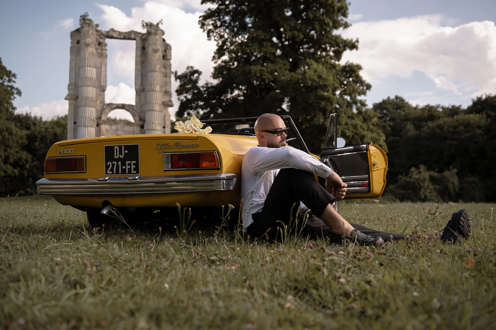 Photographer Quentin Moya sitting in the grass against a vintage yellow Alfa Romeo convertible, with stone ruins in the background