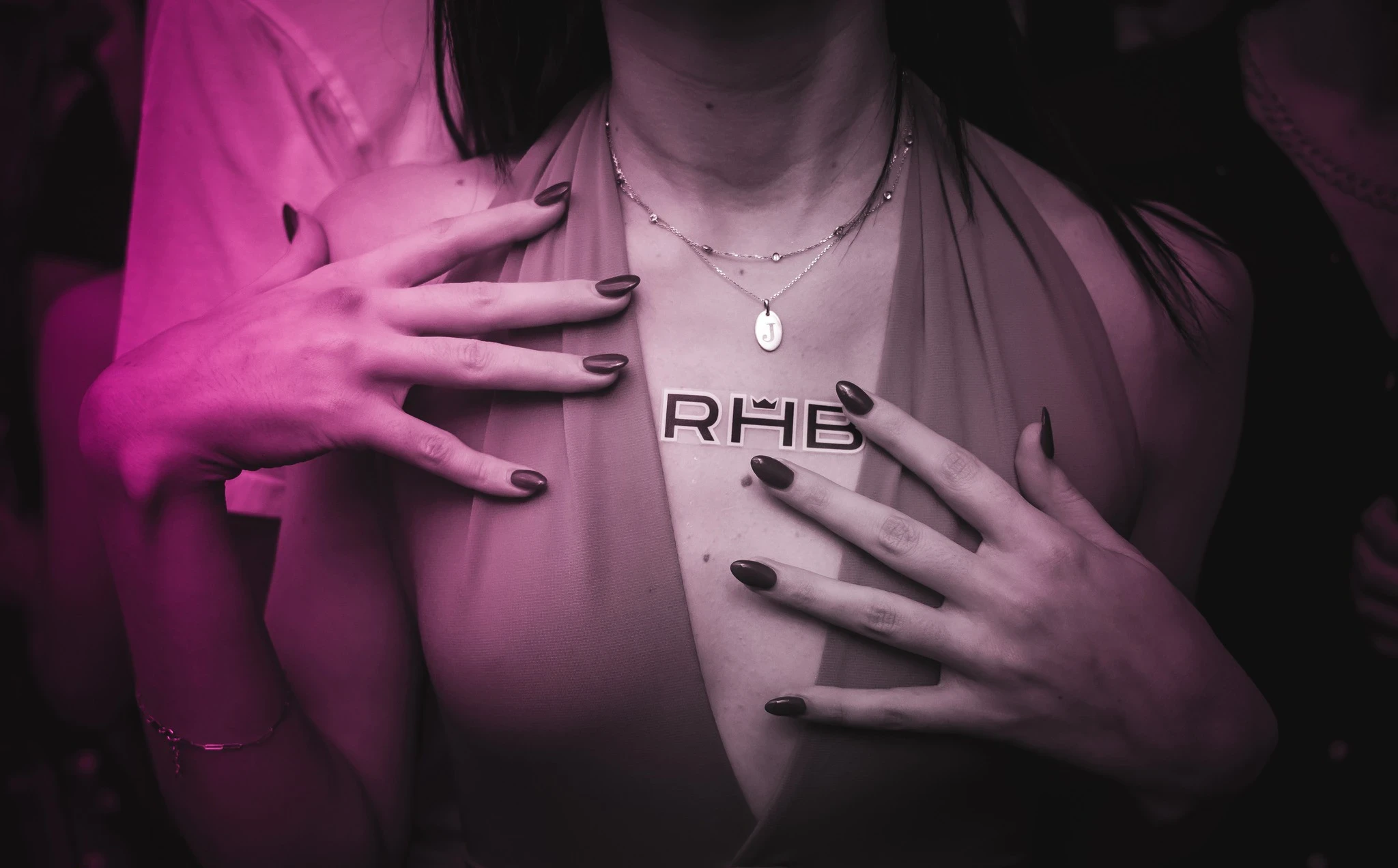 A close-up of a person's chest at the RHB party. They are wearing a halter dress and necklaces, and their hands are placed on their chest, framing an "RHB" sticker.