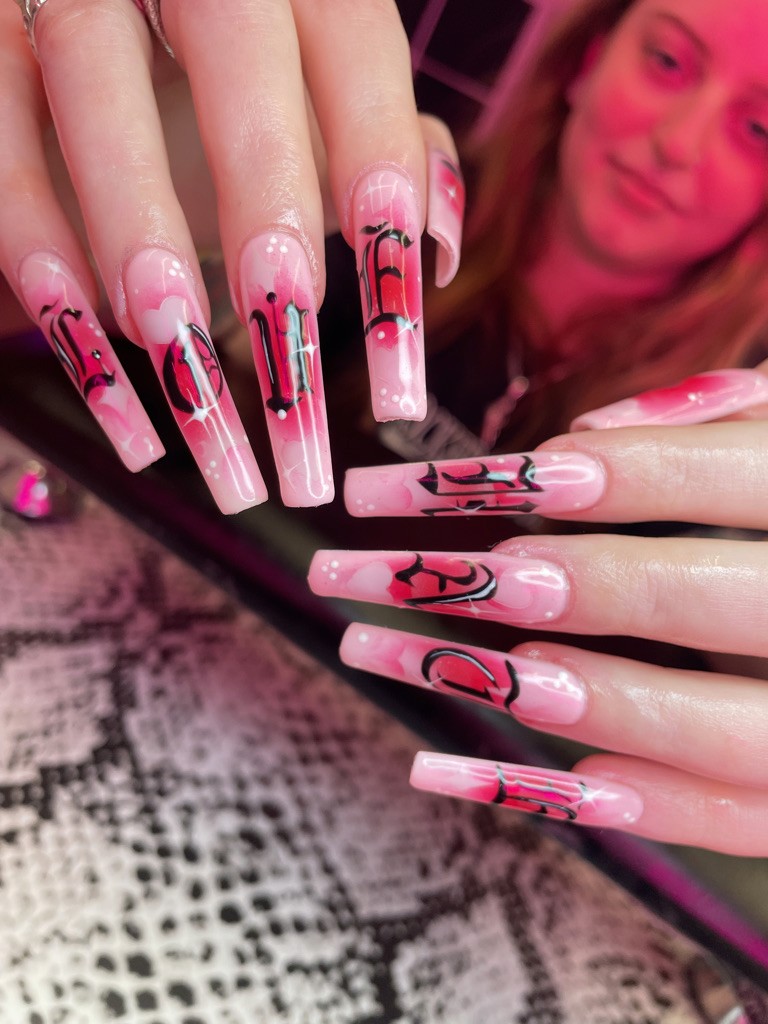 (Close-up) Extra-long square nails with a soft pink ombré and white "sparkle" details. Features bold, black Old English-style calligraphy that spans across the fingers to spell out "LOVE" and "HATE."