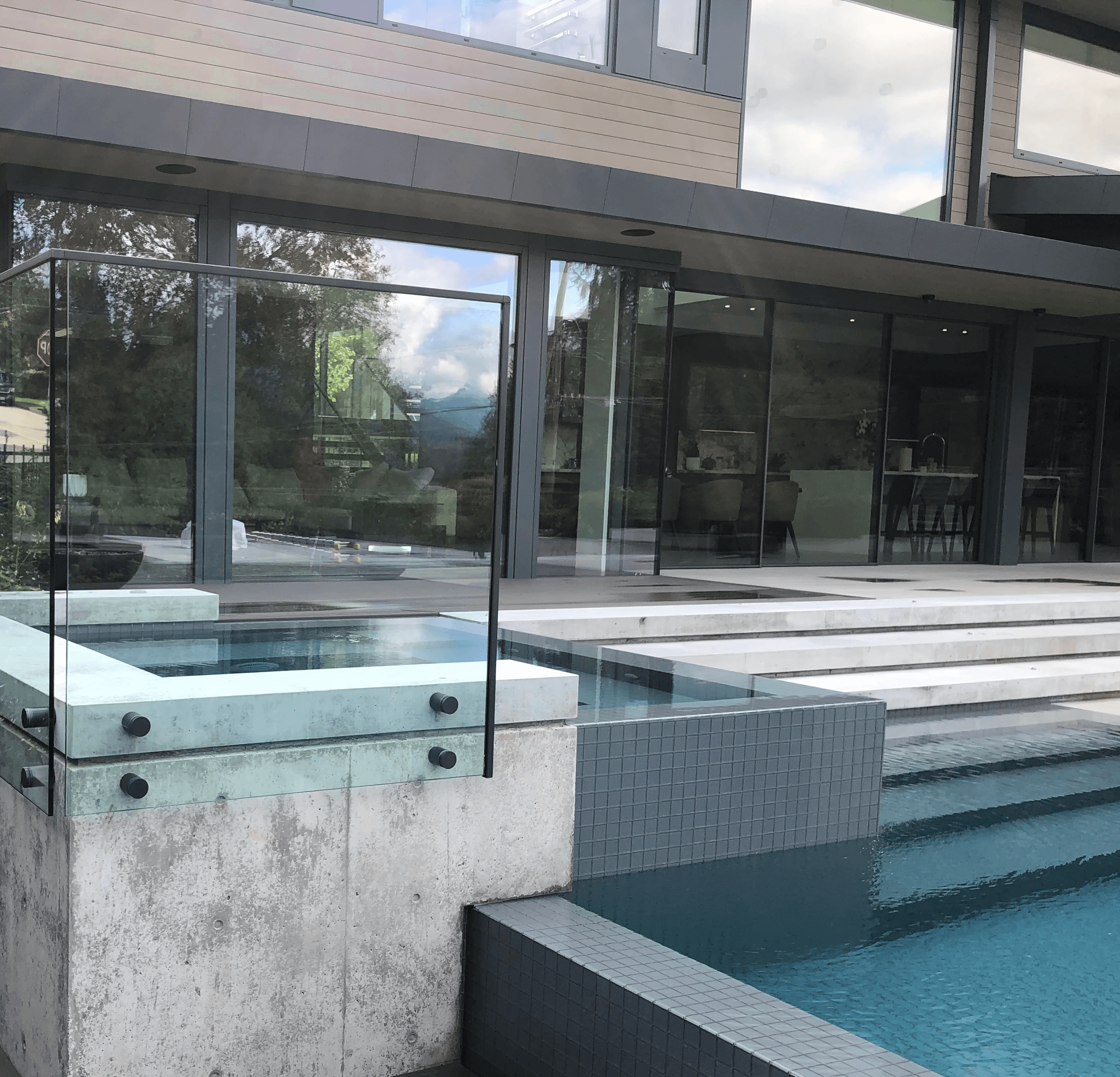 A close-up architectural shot of a luxury modern home's exterior pool deck. In the center, an elevated concrete structure contains a square hot tub enclosed by a frameless glass railing secured with black cylindrical spigots. To the right, a tiled swimming pool with clear turquoise water features wide steps leading down. The lower left shows a dark wooden plank walkway, while the large glass windows of the house reflect the sky and surrounding greenery.