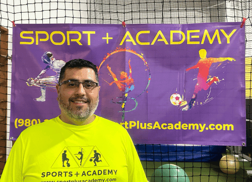 Head Soccer Coach Elio posing for a headshot in front of a purple SportPlus Academy sign hanging in our gym 