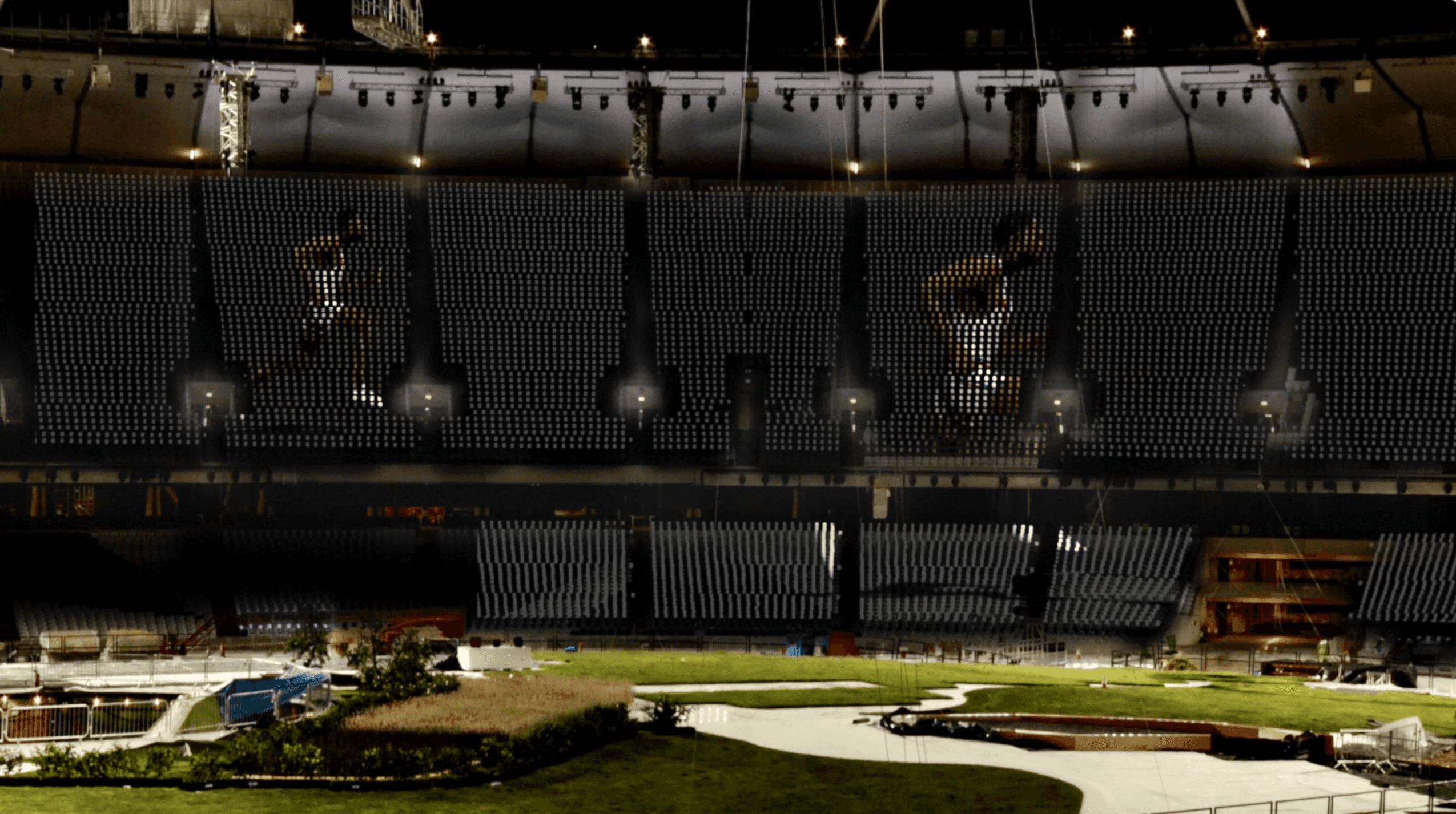 The image features a large indoor stadium under construction or preparation, with an expansive grass area and visible seating sections, illuminated by overhead lights and displaying digital projections of athletes on the walls, highlighting a blend of natural elements and digital technology.