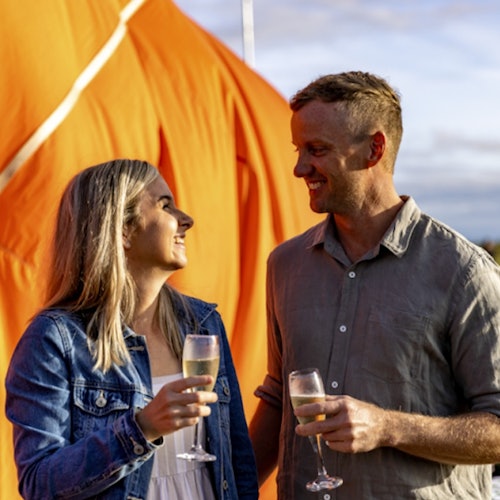 Een man en een vrouw glimlachen naar elkaar terwijl ze glazen wijn vasthouden. Een grote oranje structuur bevindt zich op de achtergrond.