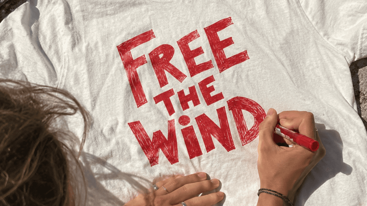 A person hand-lettering “Free the Wind” in red marker on a white T-shirt.