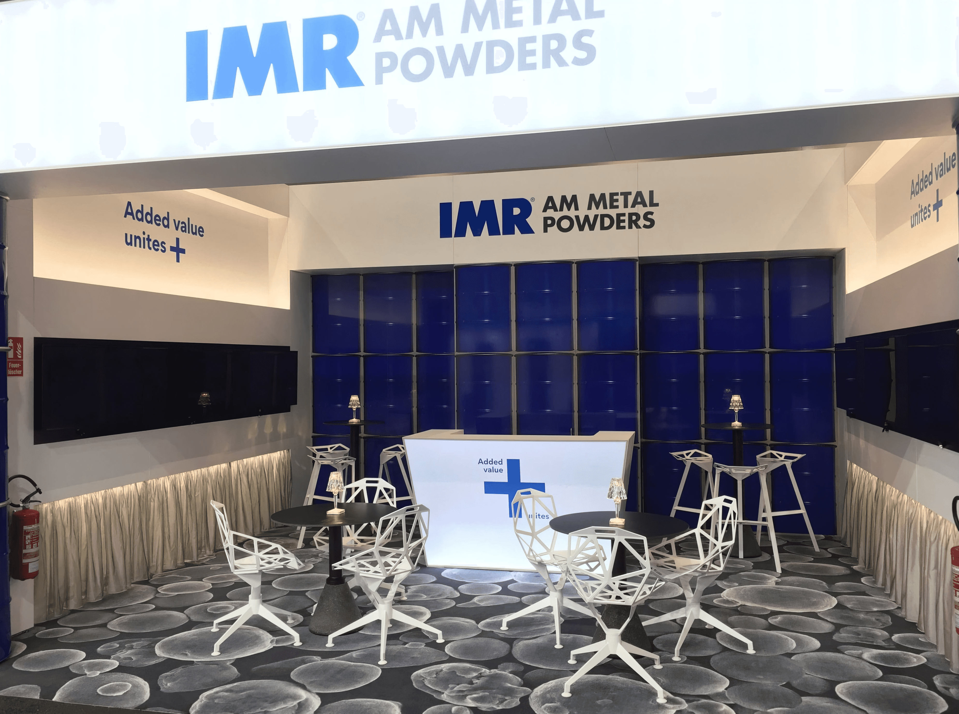 MW Interior  - Messestand IMR AM Metal Powders mit blauer Rückwand, weißem beleuchteten Tresen, geometrischen weißen Designstühlen und graphischem Bodendruck" Beschreibung: Professionell gestalteter Messestand mit leuchtendem Markenblau, geometrischen Designstühlen in Weiß und einem hinterleuchteten Empfangstresen – konsequent auf die Unternehmensidentität abgestimmte Messemöblierung für einen starken Auftritt.
