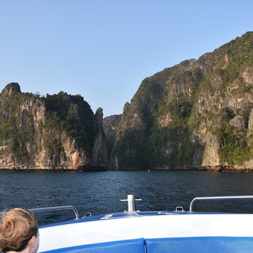 在晴朗的蓝天下，小船上的人接近被绿色植被覆盖的高大岩石峭壁。