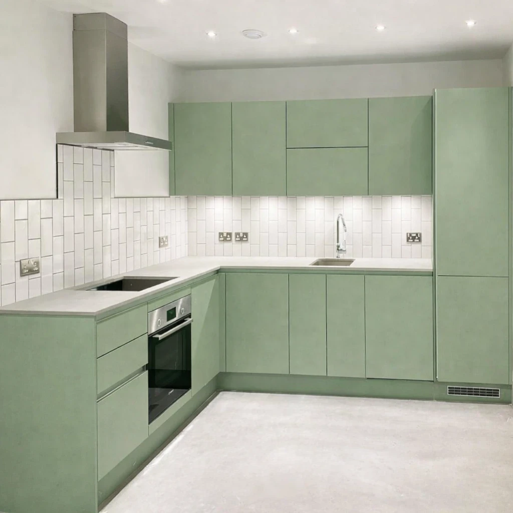 Modern L shaped kitchen with light green cabinet units, white subway tile splashback, integrated oven and hob, stainless steel extractor hood and tall fridge unit in a bright, minimalist interior.