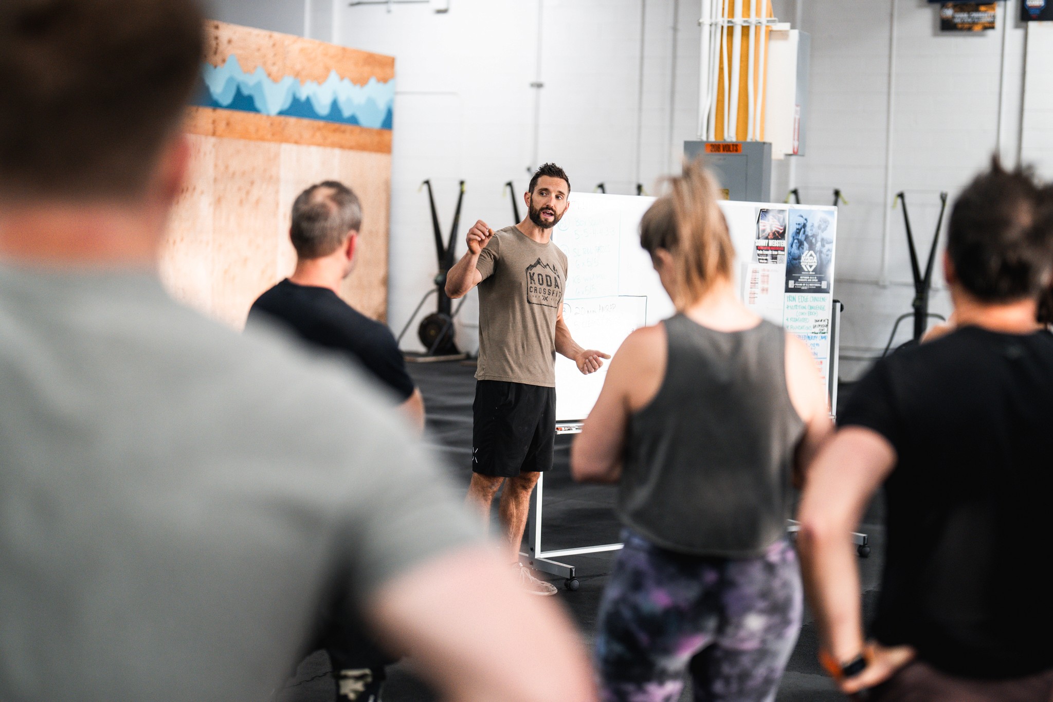 A CrossFit coach leading a small group class, with athletes listening attentively inside a brightly lit gym.