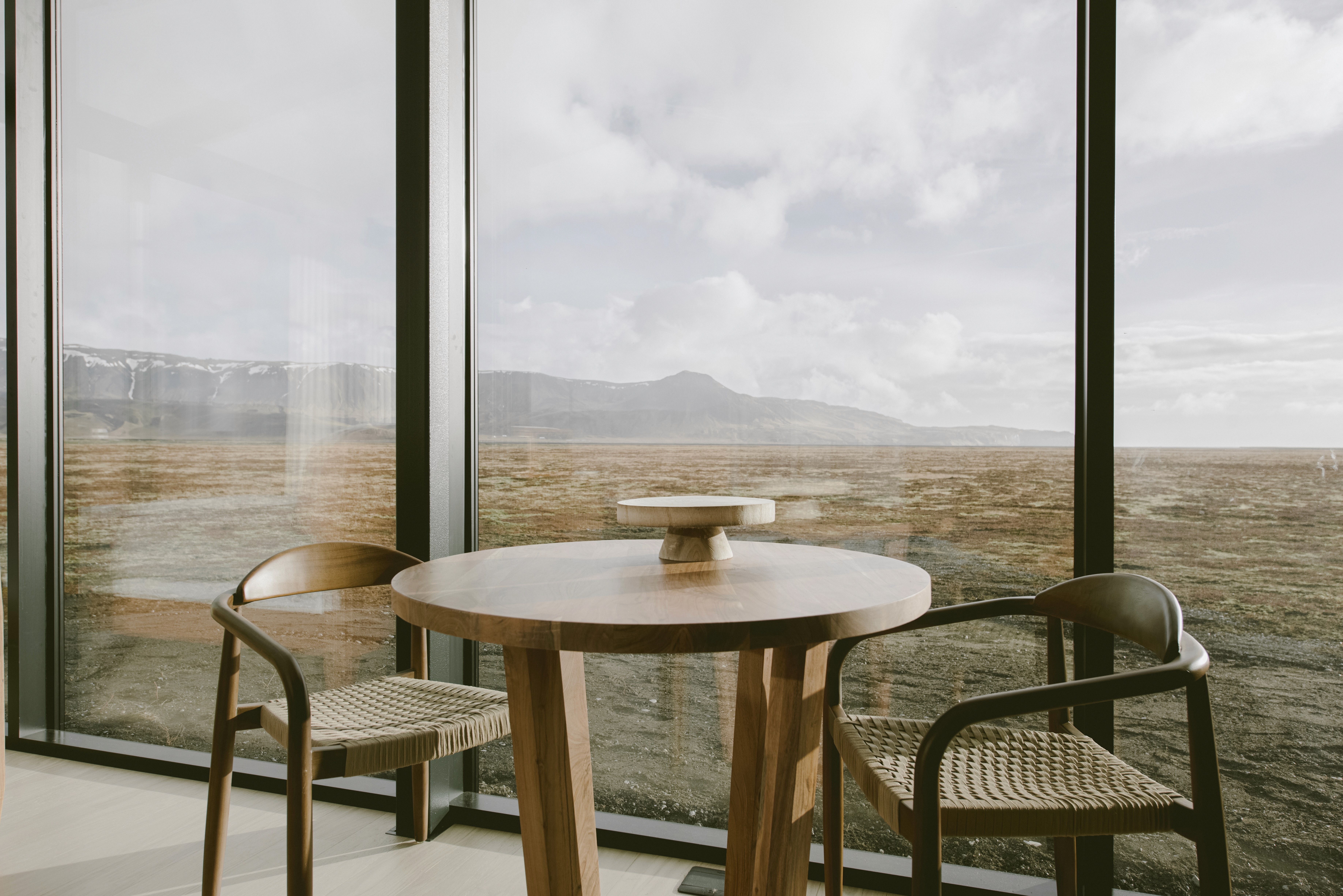 Round wooden dining table with woven chairs set by floor-to-ceiling windows, offering panoramic views of an open landscape and distant mountains from a modern design-led cabin interior.