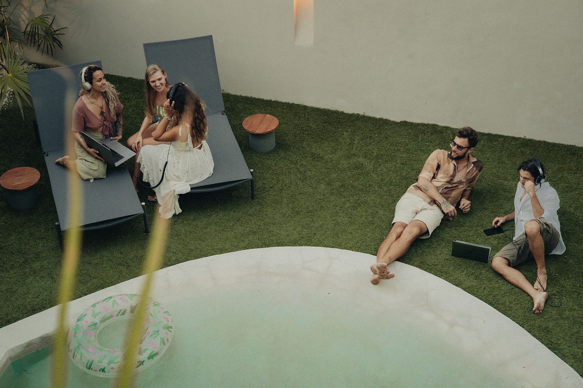 Three women sitting on sunbeds, smiling and talking while looking at a laptop, with two men nearby using a laptop and phone.
