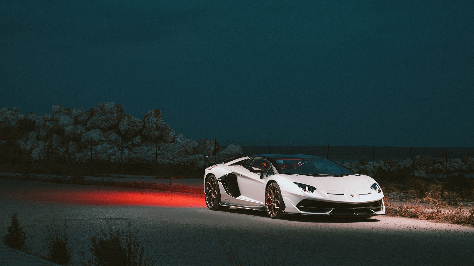 White Lamborghini Aventador SVJ with bronze wheels photographed from the front three-quarter at night, a wash of red light across the ground behind it, with a rocky coastal embankment and the dark sea on the horizon.