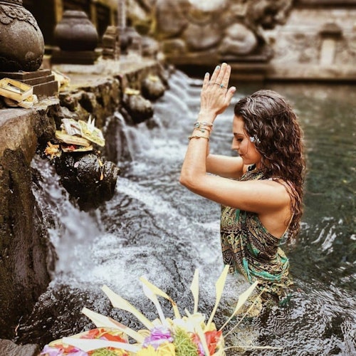 一位女性站在池塘中，雙手合十，靠近石頭噴口和供品，處於一個儀式或精神的氛圍中。