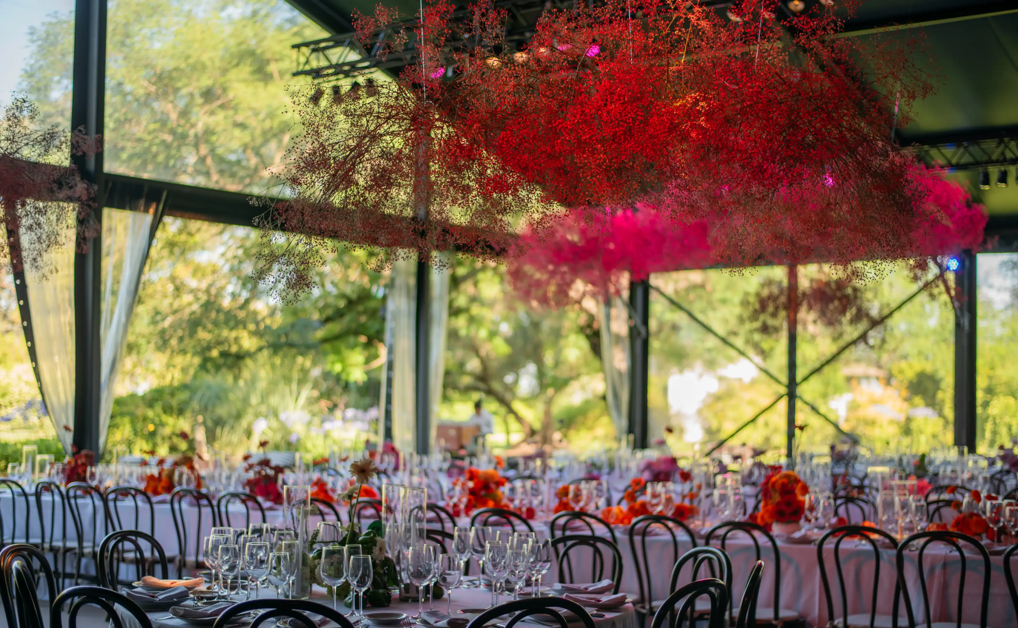Salón decorado con flores colgantes en tonos fucsia y rojo, mesas redondas con centros florales naranjas y sillas de madera oscura bajo estructura vidriada.