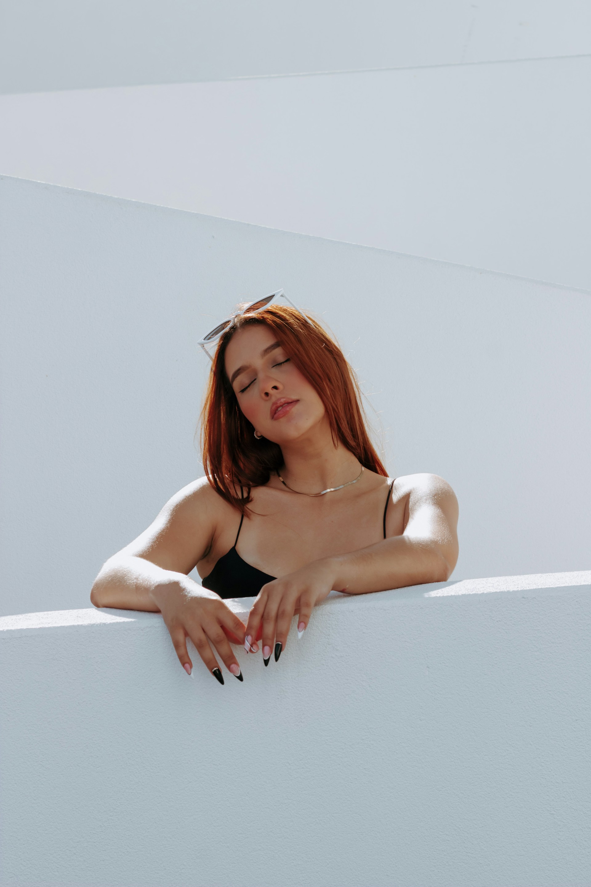 A woman with reddish hair leaning against a white architectural wall with her eyes closed.