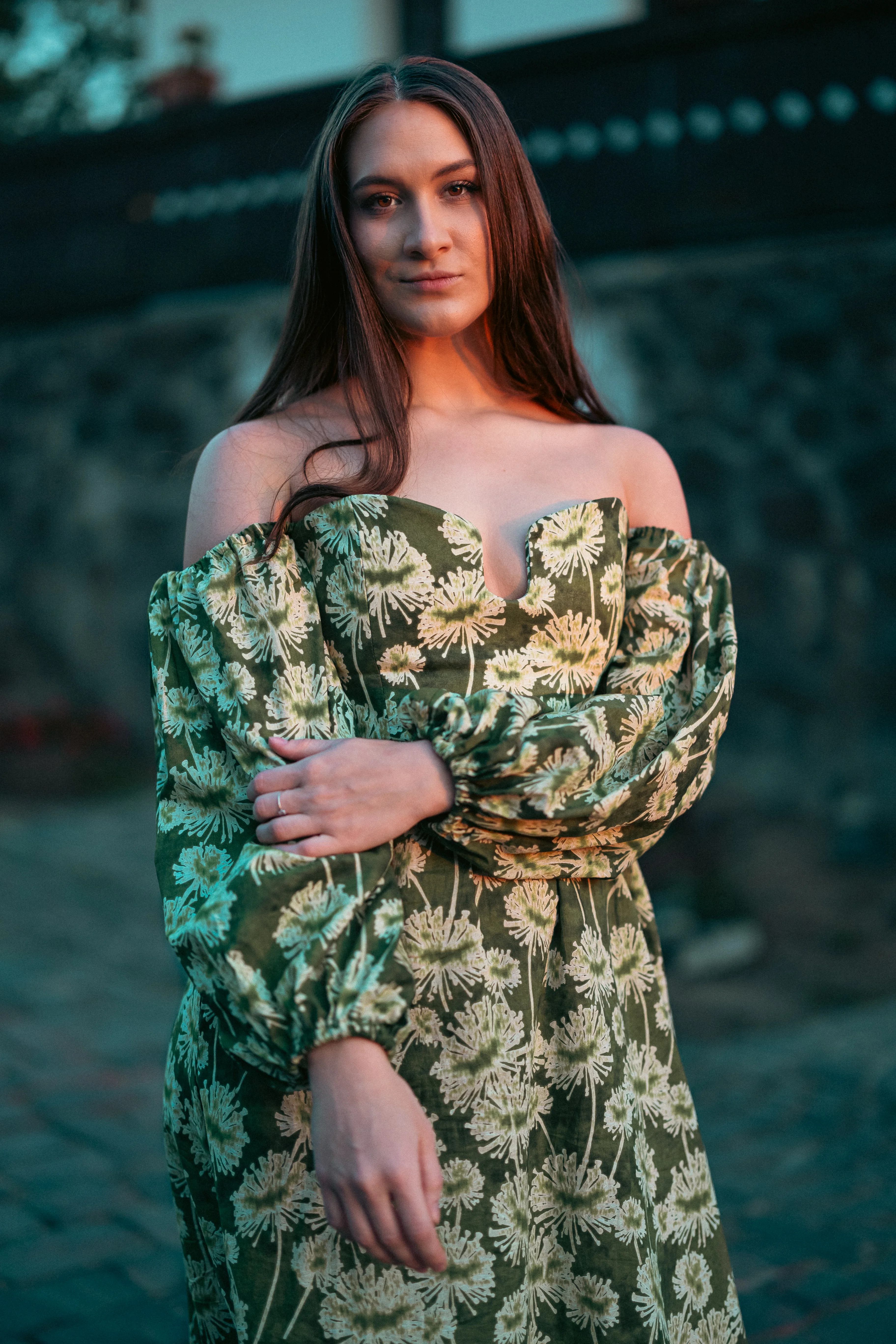 Elegáns portré hosszú hajú nőről zöld-krém virágmintás off-shoulder ruhában Hollókő történelmi ófalujának kövezett utcáján, moody teal színekkel