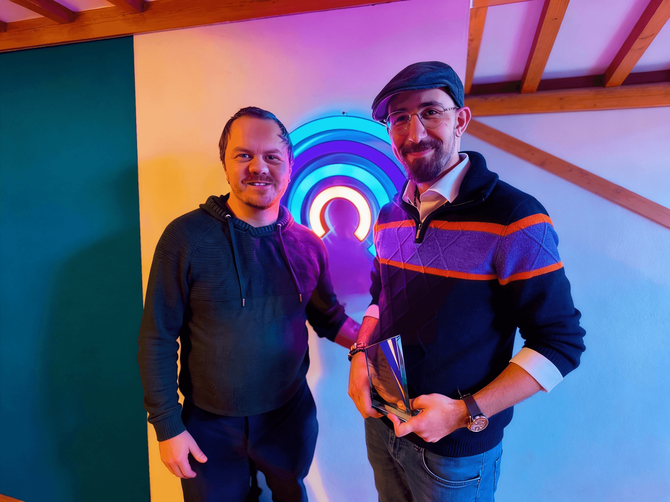 Two people standing indoors in front of a colorful neon CAMSOL logo. They are smiling at the camera; the person on the right is holding an award trophy. The lighting is warm and vibrant.