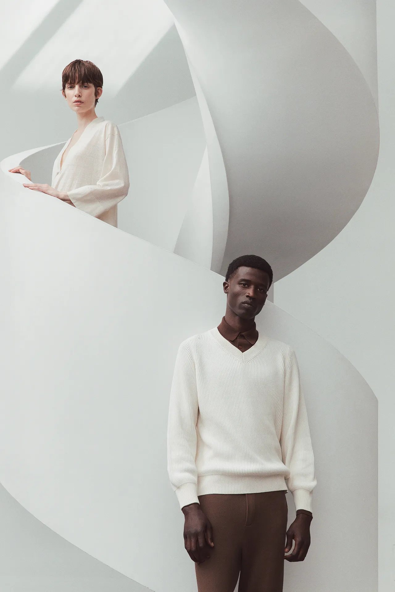 Aida Barni SS26 collection campaign featuring a male model in a cream ribbed V-neck sweater and a female model in a white knit top on a modern sculptural staircase.