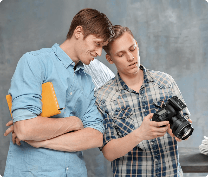 A person in a white shirt holds a DSLR camera up to their face, taking a photo. The background is blurred, giving the image a focused and artistic tone.