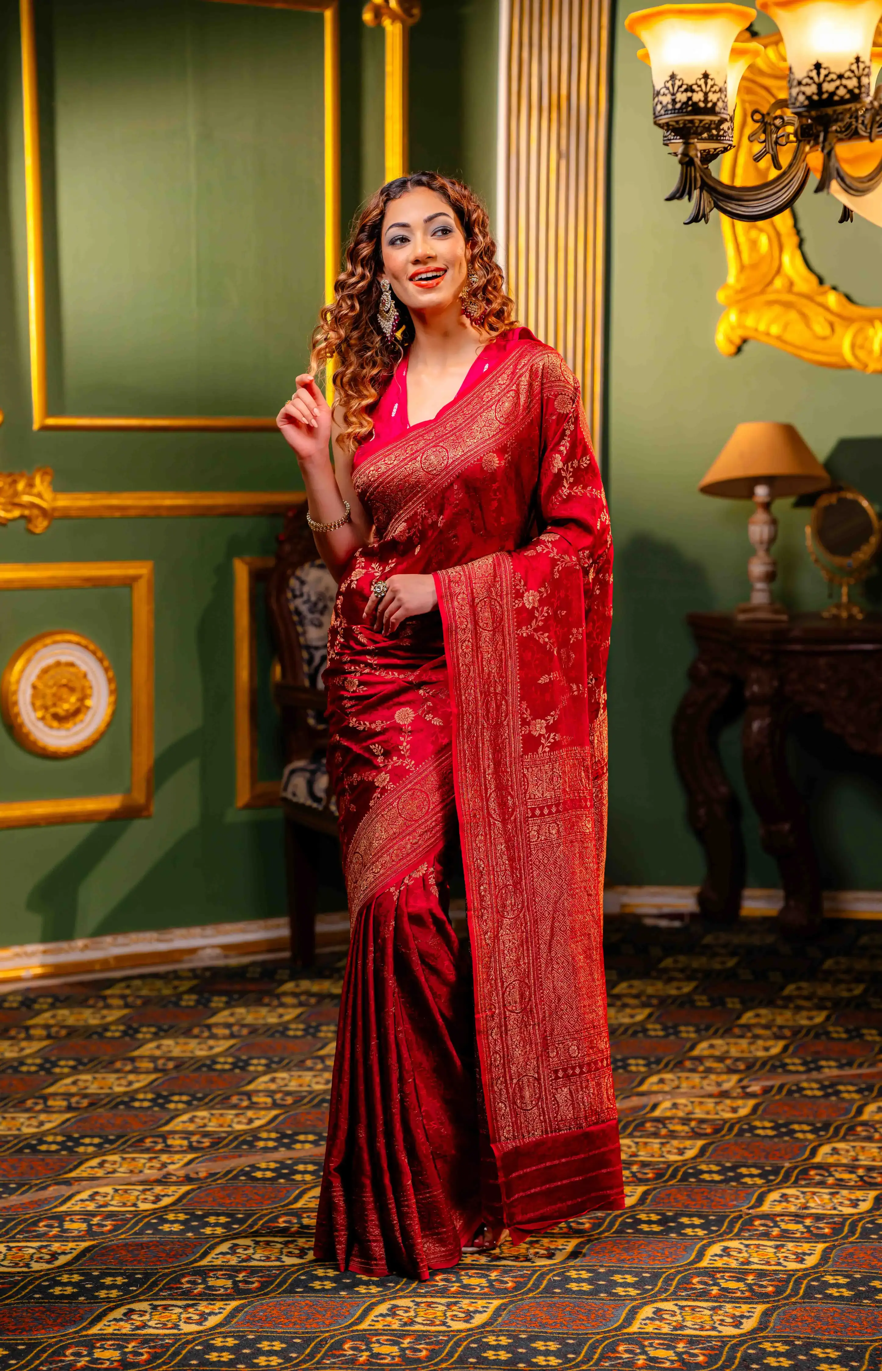 A woman wearing a red saree with golden borders from That Saree Shop, standing gracefully in an elegant room, highlighting the saree’s luxurious weave and classic festive charm.
