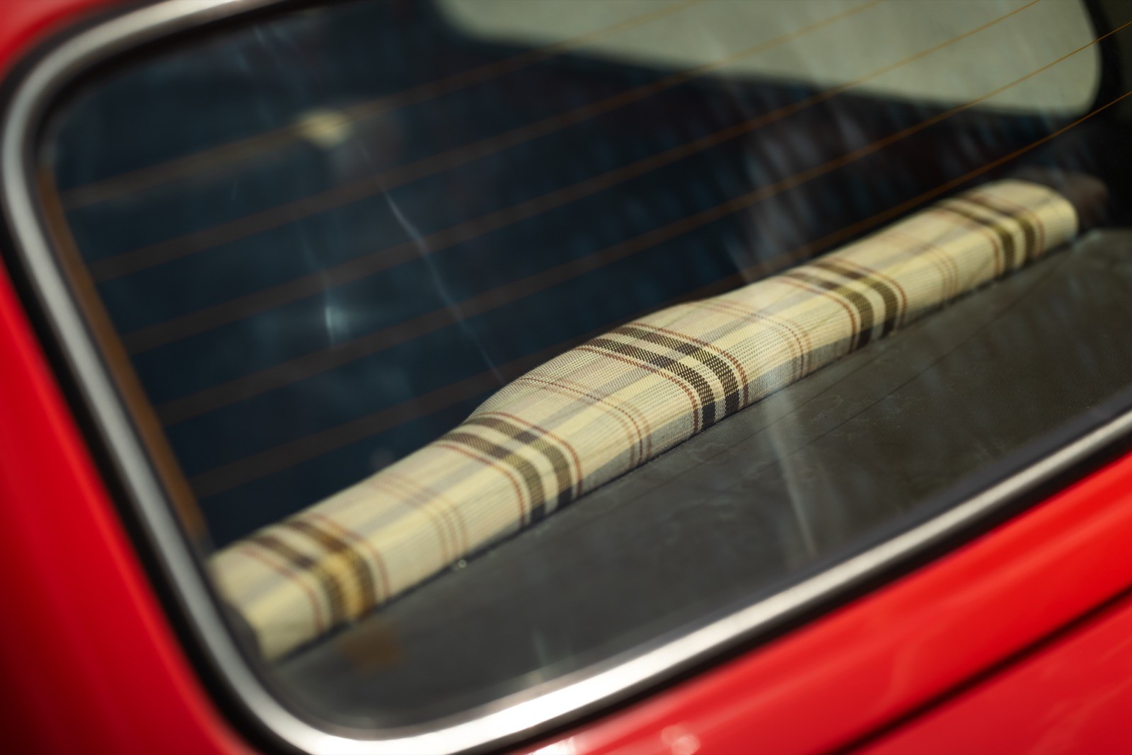 Window view of a vintage car showing a rolled-up beige blanket on the back seat against a dark interior backdrop.
