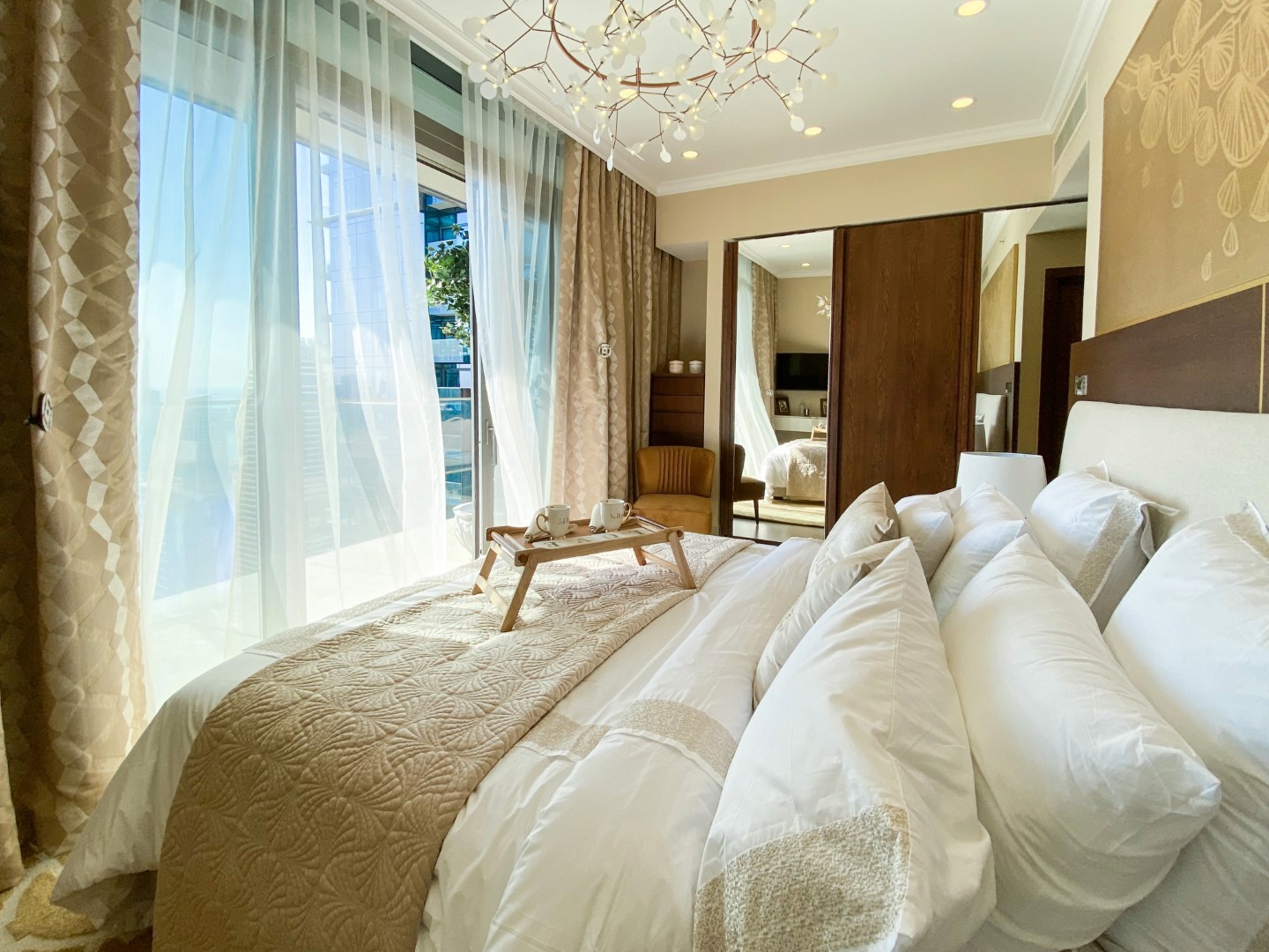 A luxurious hotel room features a plush, neatly made bed with beige and white pillows, a light brown textured comforter, a wooden breakfast tray with cups, floor-to-ceiling windows with sheer curtains, and a large mirror reflecting elegant decor, enhancing the serene ambiance.