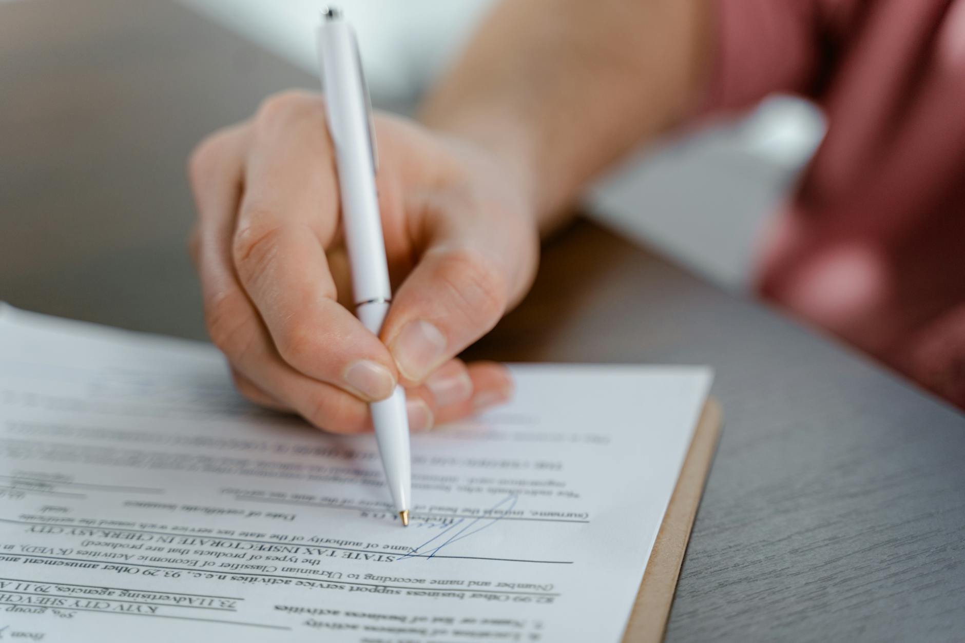 Close-up of a hand signing a printed legal document with a white ballpoint pen on a grey desk