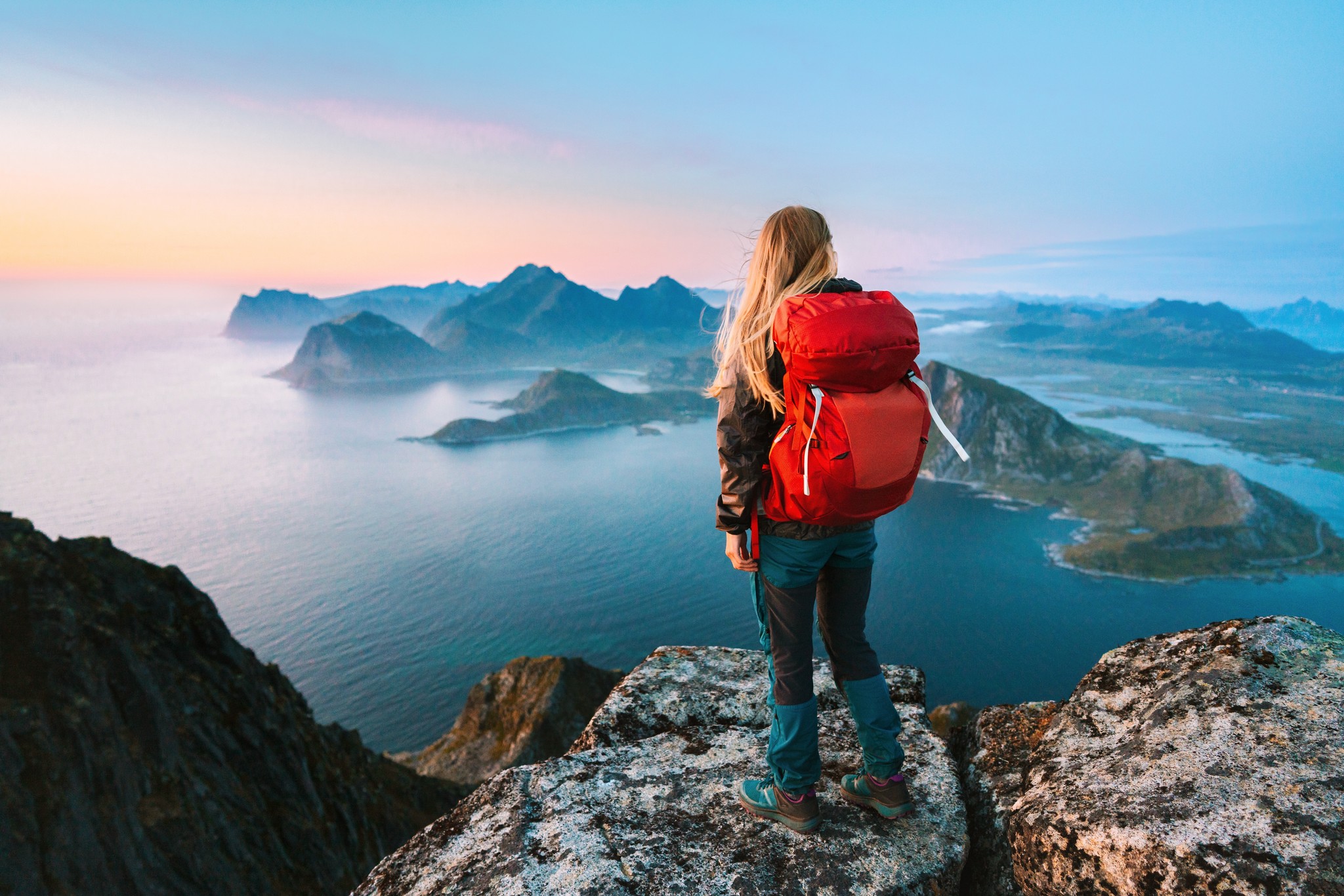 A woman in hiking books and a red backpack atop a cliff looking over a dramatic archipelago in Norway